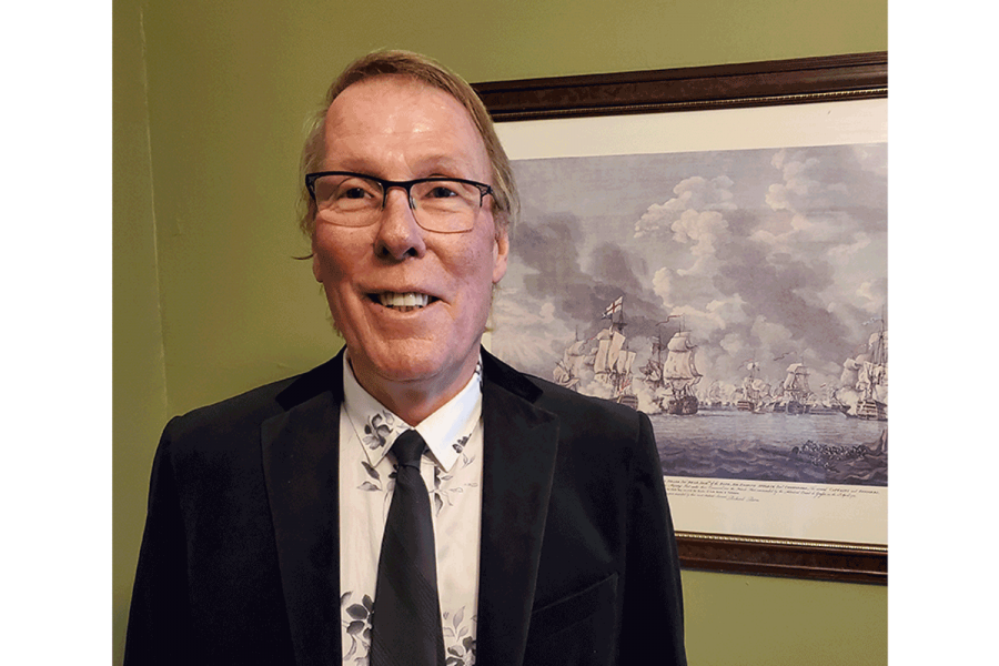 Headshot of James Fergusson standing in front of a painting of ships hanging on a green wall.