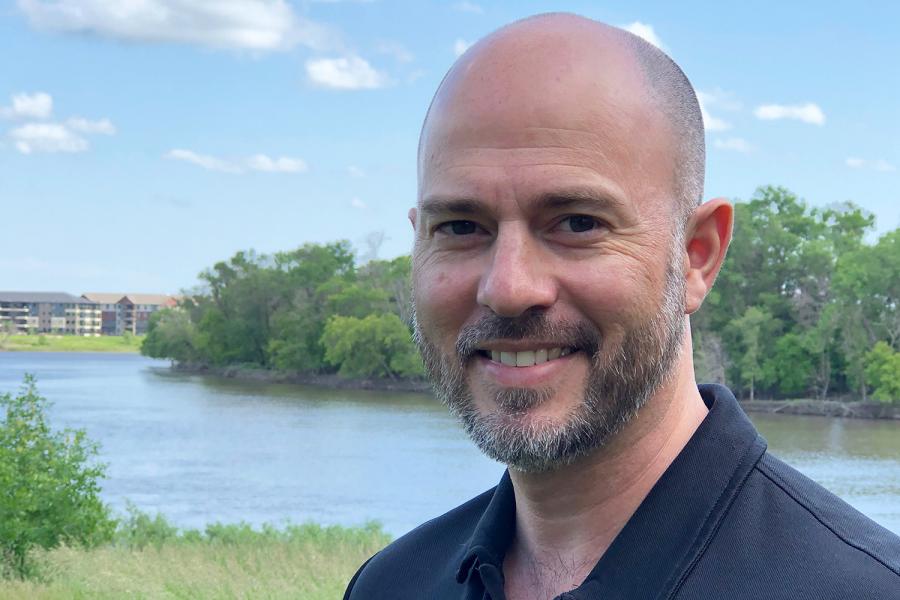 Dr. Shawn Clark smiling and posing for the camera outside on a bright sunny day. Wearing a navy blue polo with facial hair and a bald head. In the background is a river with condos in along one edge and trees on the other. 