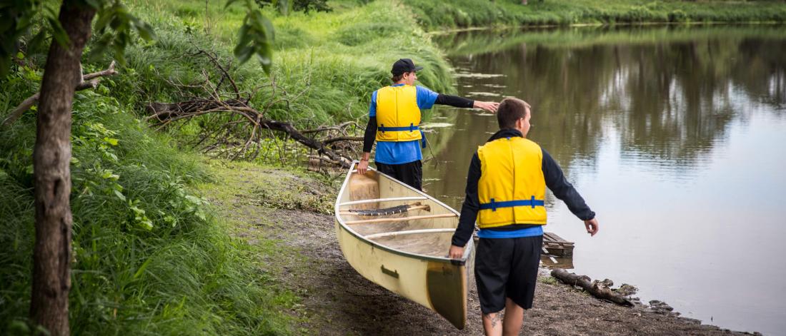 Two recreation management and community development students carry a canoe along the shore of a river.