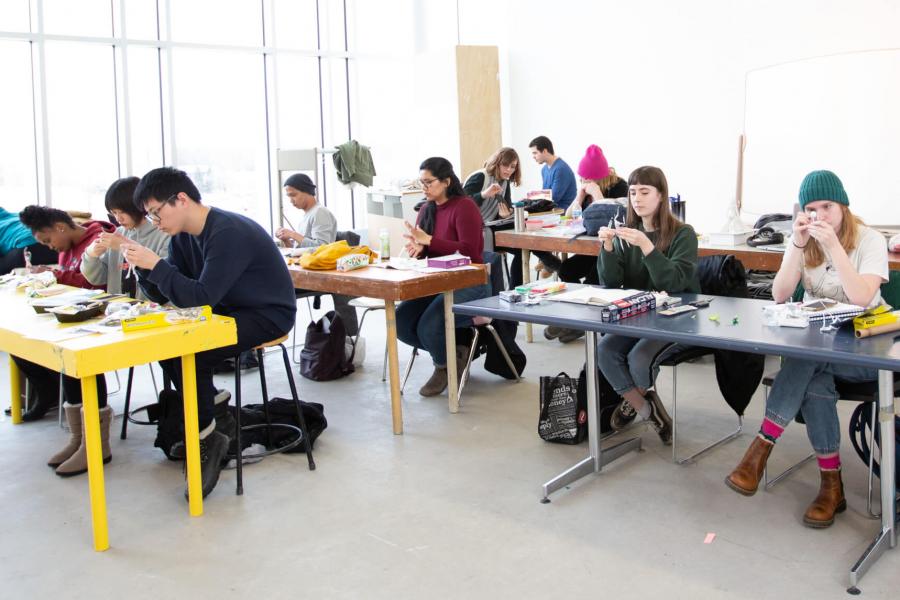 A class of first year school of art students work in a studio at long narrow tables.