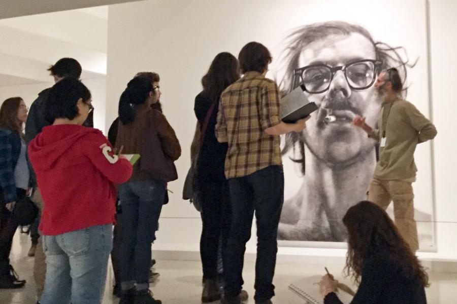 A man stands in front of a piece of artwork, students gather around and listen to him speak about the piece.
