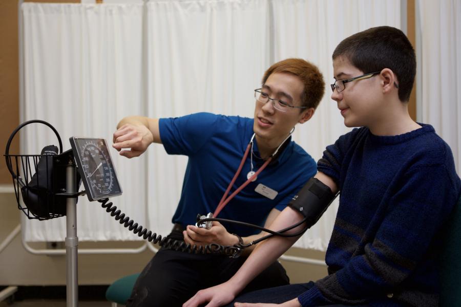 A rehabilitation sciences student uses a blood pressure monitor on a young patient and points to the monitor to show how it works.