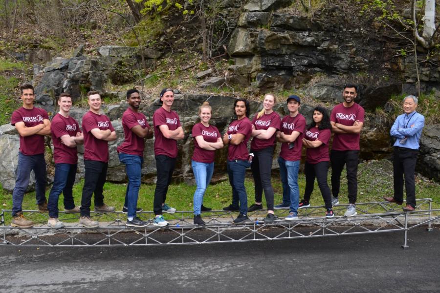 A group of 11 civil engineering students and their faculty advisor stand arms crossed atop a bridge like structure they have built.