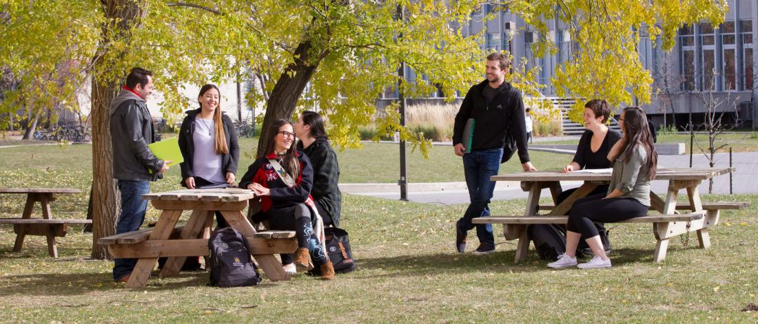Seven Indigenous students sit and stand together outdoors at two picnic tables at the Fort Garry campus. 