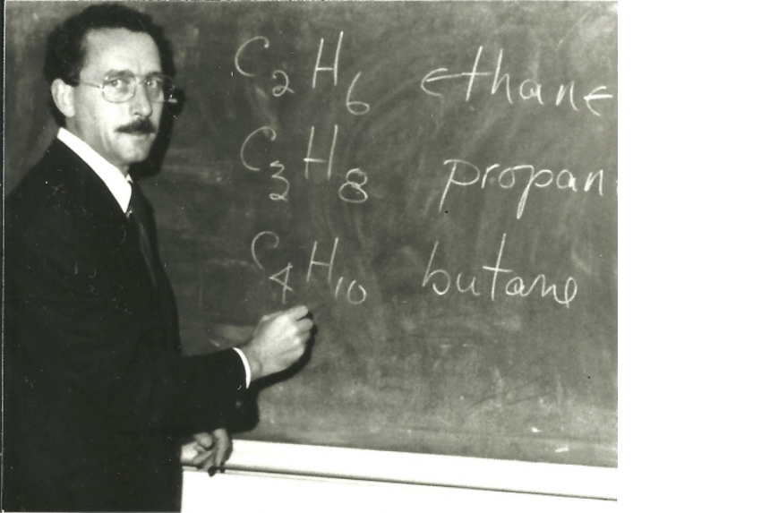 Charles Burchill looking at the camera while writing on the blackboard. The photo is in sepia color.