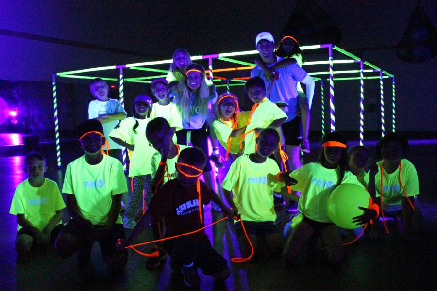 A group of Mini U juniors and their leaders posing for a group photo in a dark room with neon glowing t-shirts.