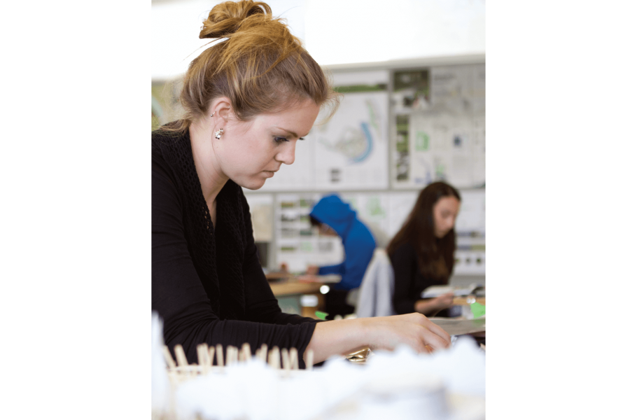 A students works on a project at a desk. Several other students can be seen working in the background.