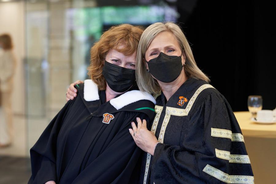 Chancellor Mahon stands with her arm around Laurel Hyde, Chair of the UM board of governors. Both are wearing academic gowns and covid masks. 