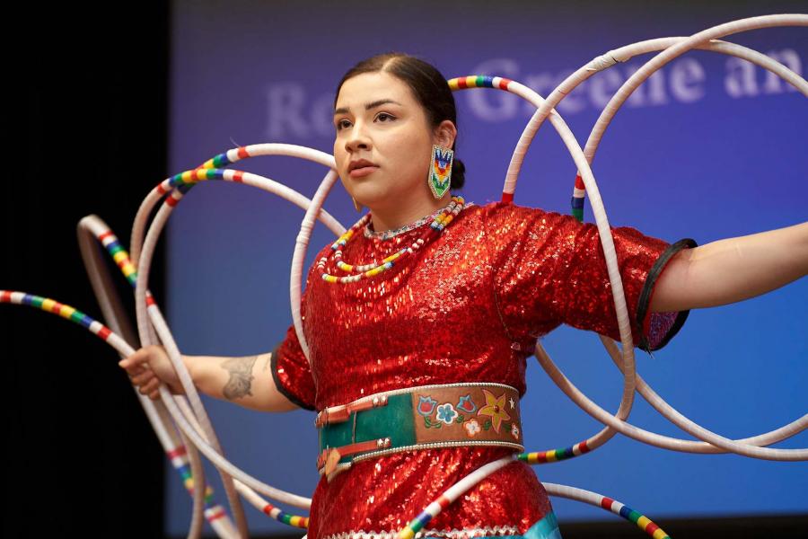 An Indigenous woman wearing red regalia and beaded earrings dances with her arms outstretched intertwined with several hoops.