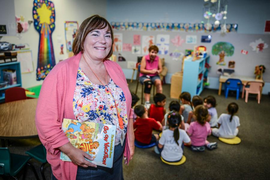A smiling play care instructor wearing a pink cardigan holds a Berenstain Bears book in front of a brightly decorated room where children are sitting on the floor in front of another instructor.