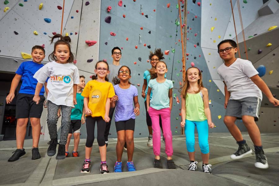 A large group of children jump up and down with excitement in front of a climbing wall.