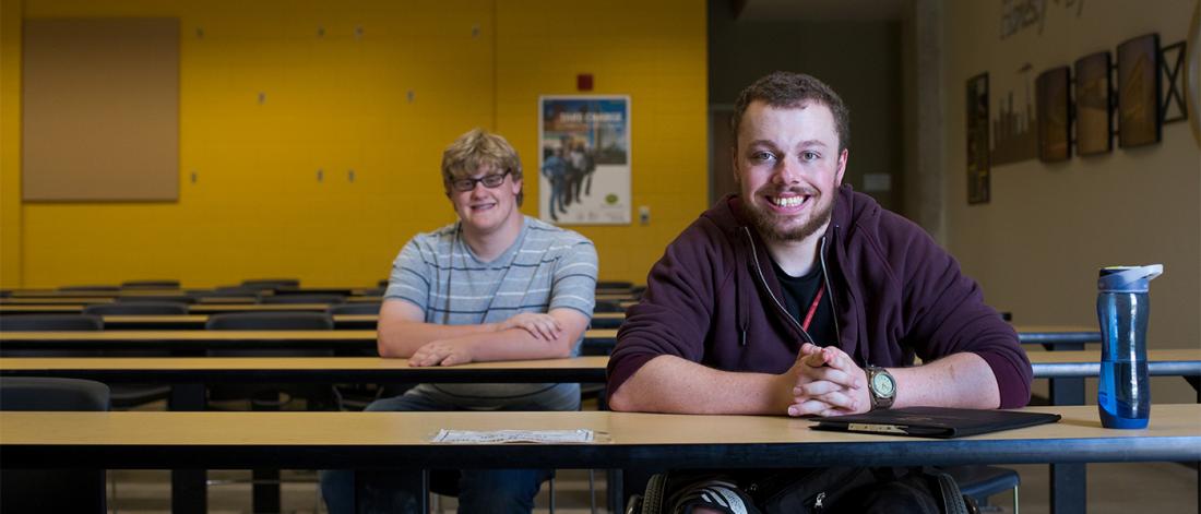 Two students sitting in class looking at the camera one of them is on a wheelchair.