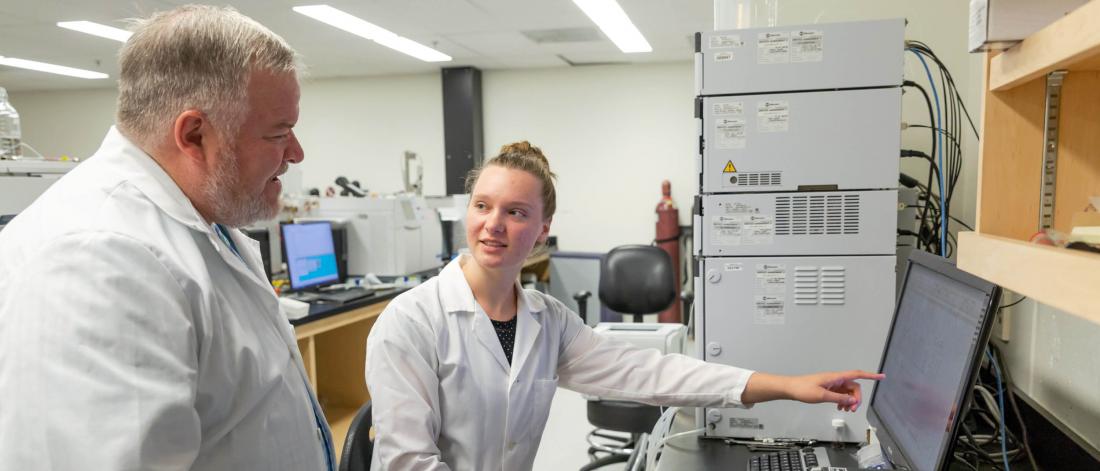 A student points to a computer monitor while talking with a professor in a research lab. 