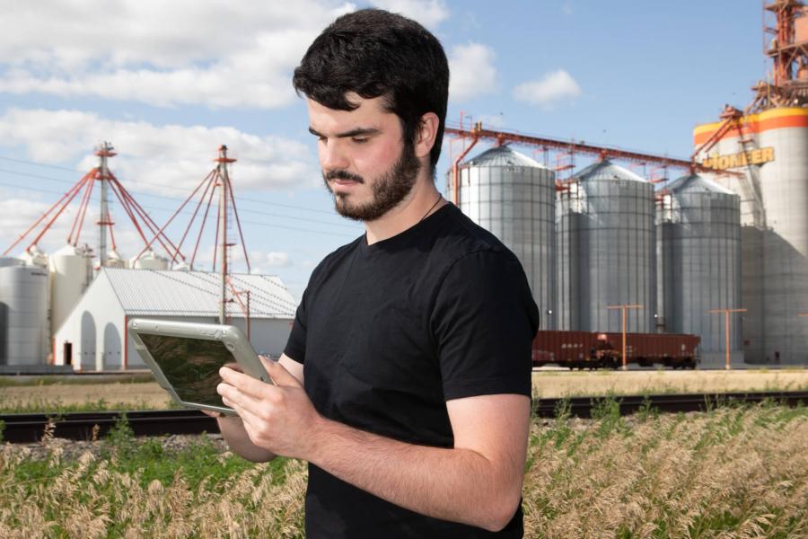 A student uses a piece of equipment while standing in a field with a large grain elevator in the background.