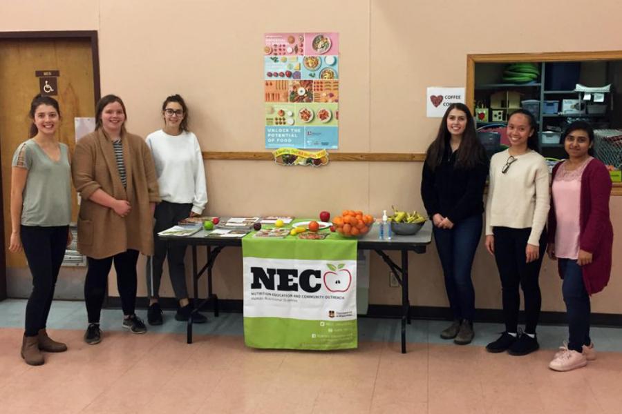 A group of six students stand on either side of a table with various fruit and some brochures for a nutrition education and community outreach program.