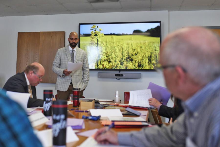 People gather around a boardroom table while someone makes a presentation, a television in the background shows a field of canola.