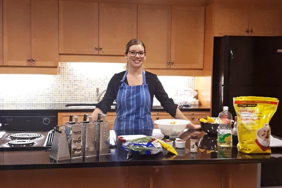 A student stands in a large classroom kitchen surrounded by ingredients and cooking supplies. 