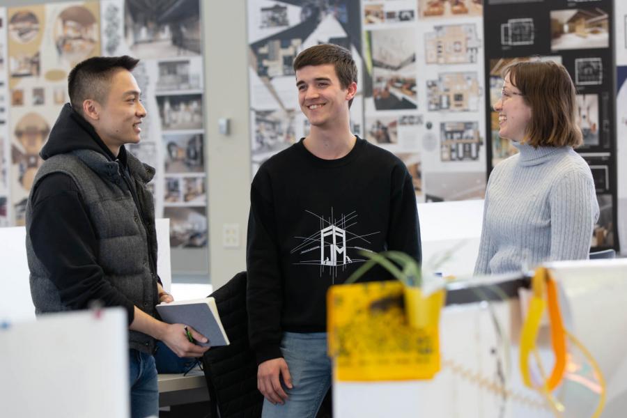 Three students from the Faculty of Architecture standing in a classroom talking to each other.