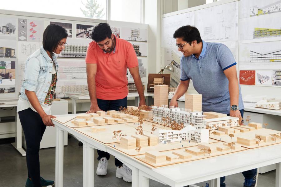 Three architecture students gather around and discuss an architectural model of a city.
