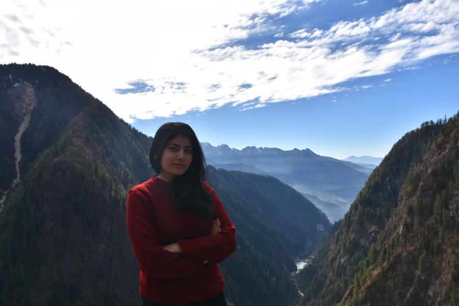 Person in a red sweater standing in front of a mountainous landscape with a bright sky and scattered clouds.
