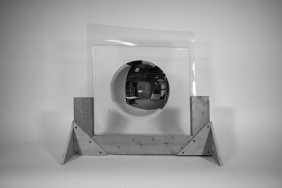The art piece features a convex, reflective half-sphere made of C.A.S.T. plastic, protruding from a textured square backdrop. It is enclosed in a wooden frame with triangular side supports, emphasizing its geometric form and reflections.