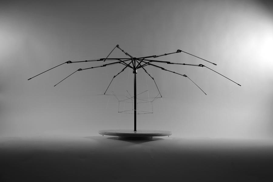The image showcases a construction resembling an umbrella frame, with slender metal rods extending symmetrically from a central point. Fishing lines and wires create geometric patterns, adding complexity to the structure, which is softly lit against a gray background.