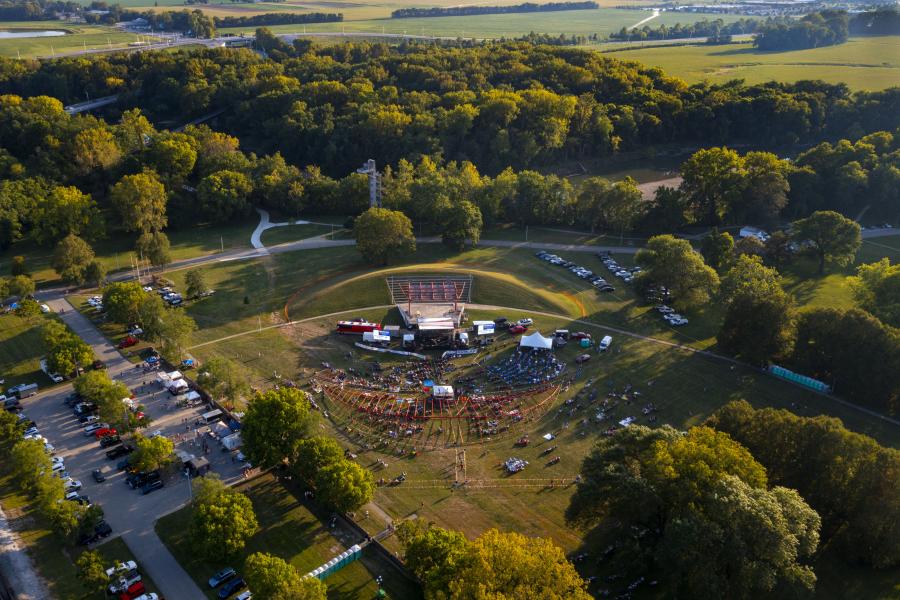 Aerial view of an outdoor amphitheater with an event taking place, surrounded by forest and fields.