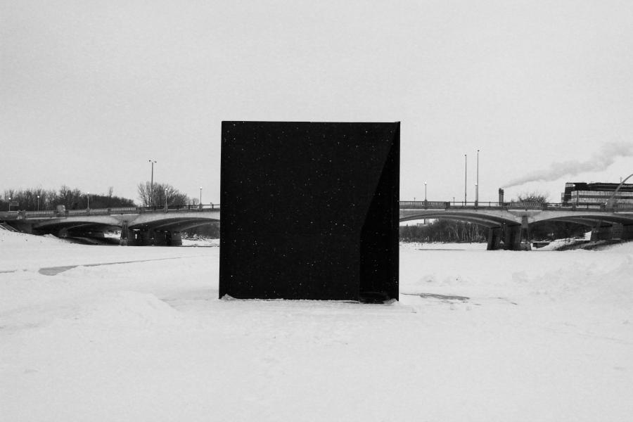 A large black monolith stands in a snowy landscape with a bridge in the background.