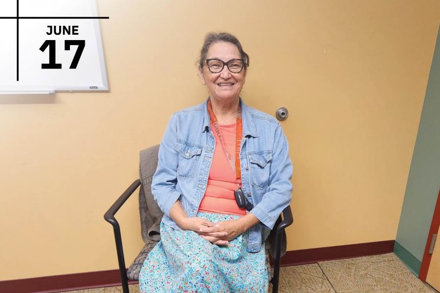 Grandmothers sitting on an office chair smiles infront of the camera as she wears a bright blue skirt and denim button up top.