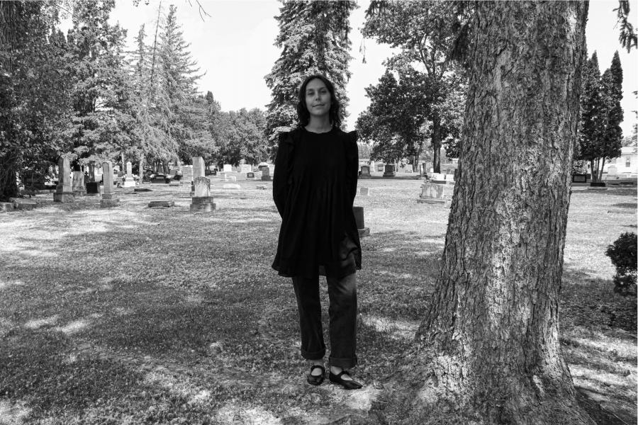 A woman in a black dress and jeans stands beside a tree in a cemetery, surrounded by tombstones and trees, conveying a serene and contemplative mood.