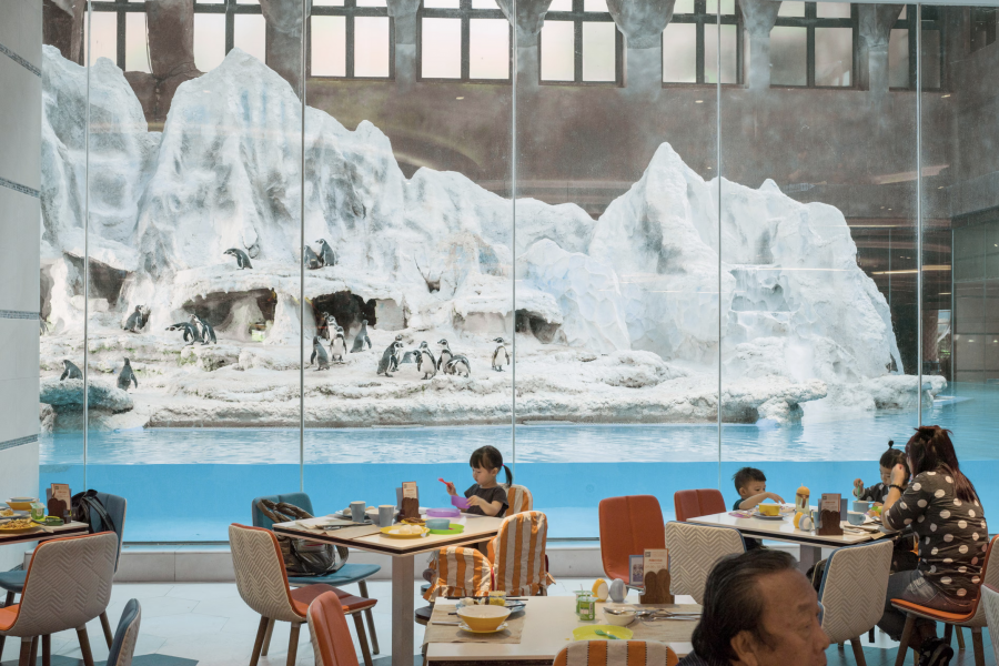 People dine in a restaurant with large windows displaying a snowy penguin habitat outside. The scene blends indoor dining with an icy landscape.