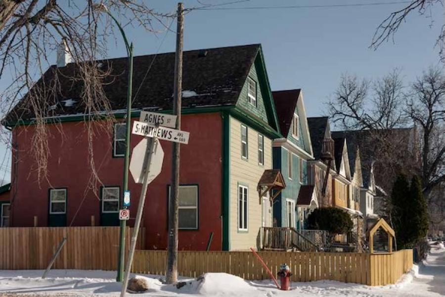 Row of colorful houses with peaked roofs under a clear blue sky. Snowy street corner with a wooden fence and bare trees. Street signs read "Agnes St" and "St. Matthews Ave."