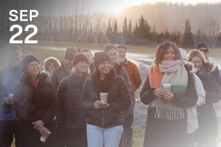 A group of people in winter clothing, smiling and holding cups, stands outdoors at sunset.