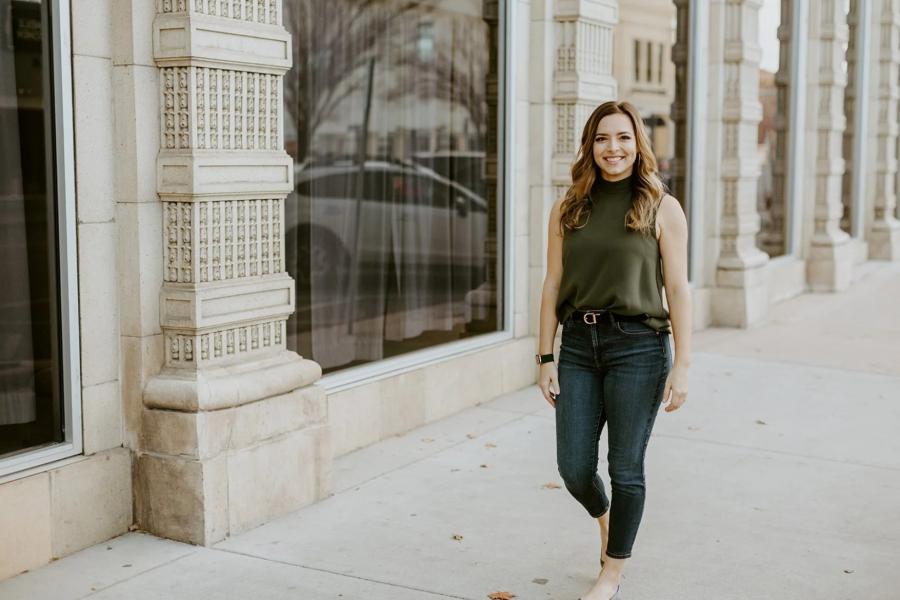 A woman in a green sleeveless top and jeans walks confidently on a city sidewalk. The architecture features large windows and ornate stone columns.