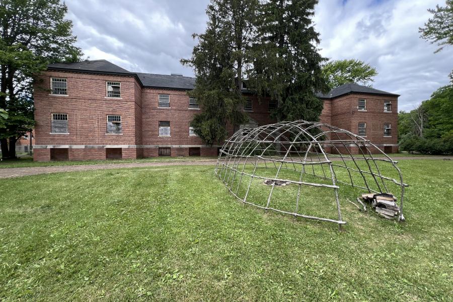 A large, old red-brick building with small windows stands surrounded by trees. In front, a dome-shaped metal sculpture sits on a green lawn under a cloudy sky.