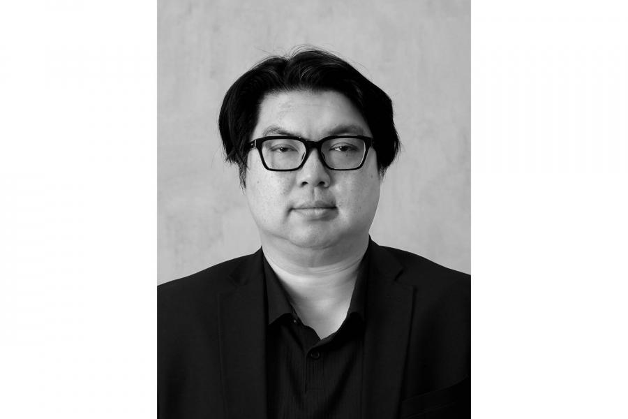 Black and white portrait of a person with glasses and dark hair, wearing a suit. They have a neutral expression against a plain background.