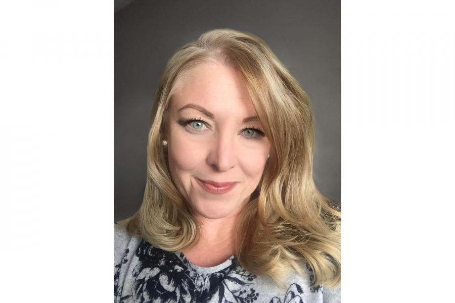 Smiling woman with blonde hair and blue eyes, wearing a patterned top against a neutral background. The tone is warm and friendly.