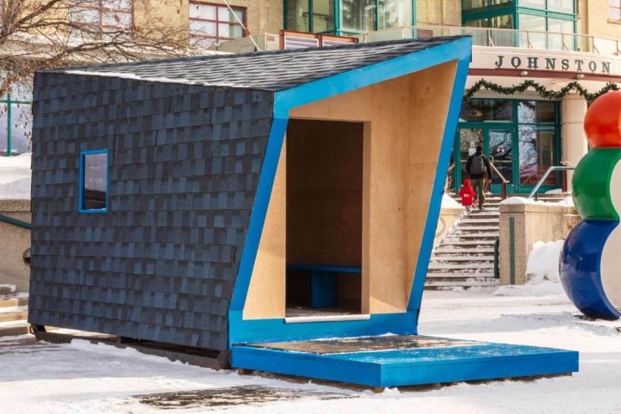 A modern, angular wooden shelter with a blue border sits on a snowy sidewalk. A colorful sculpture and steps leading to a building are in the background.