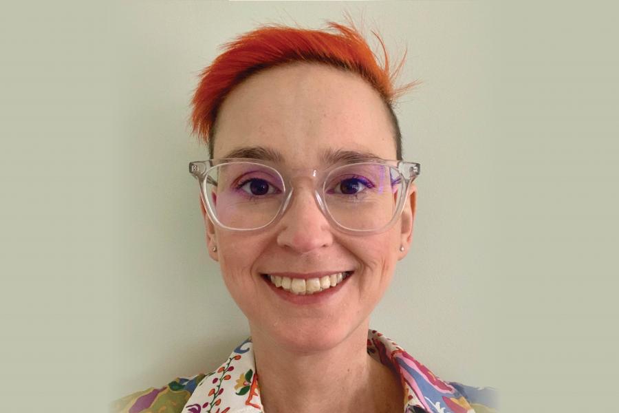 A person with short, bright red hair and clear glasses smiles warmly. They're wearing a colorful, patterned shirt, exuding a cheerful, friendly vibe.