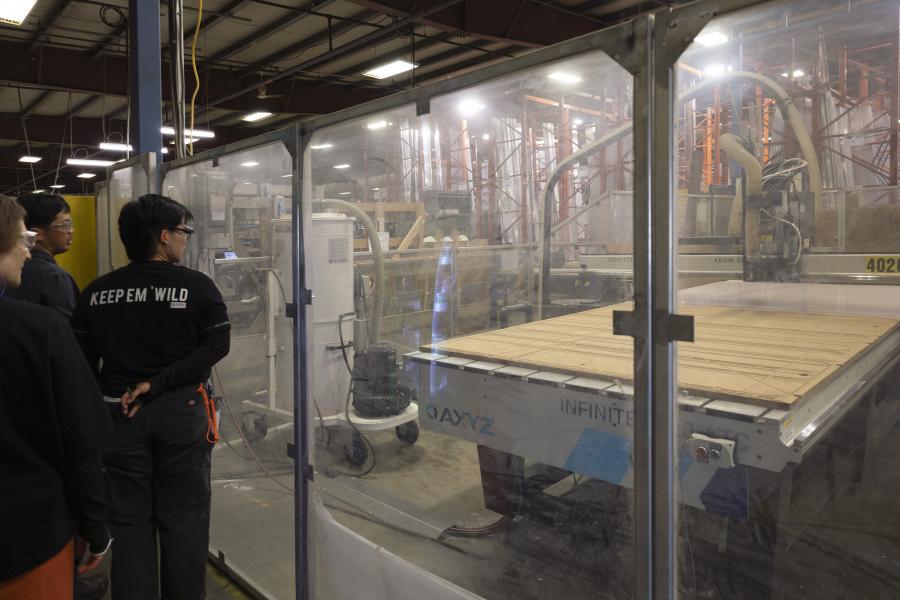 Group of people in safety gear observe a large CNC machine through glass in a factory. The scene conveys a sense of industrial innovation and safety.