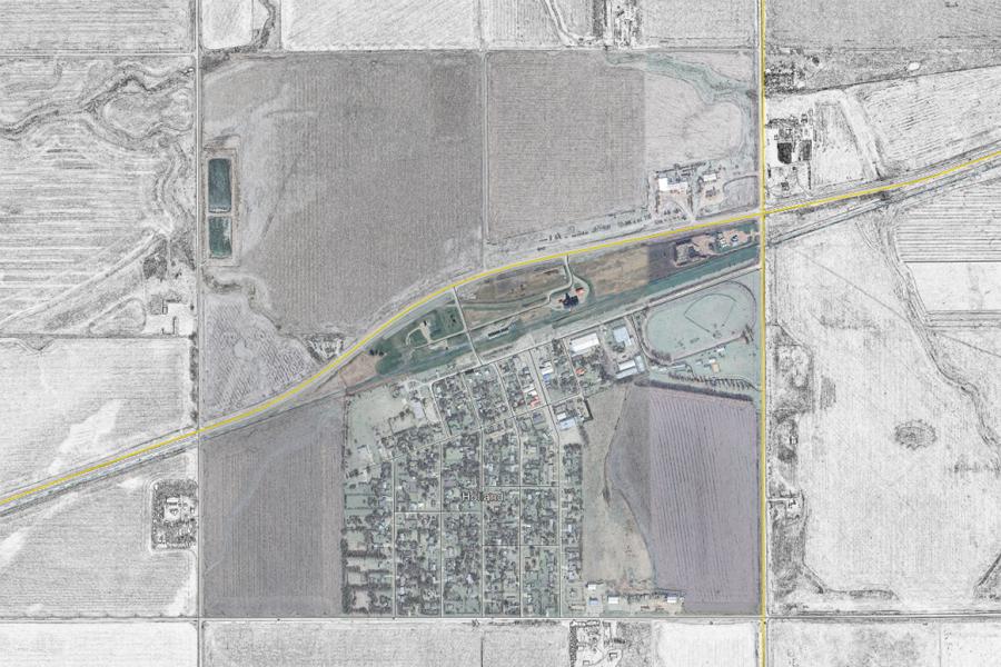 Aerial view of a rural town surrounded by fields. A grid of streets and houses is in the center, with roads crossing the top and right sides. The tone is neutral.