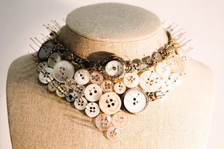 A creative necklace made of white buttons, watch faces, and safety pins is displayed on a beige mannequin bust, evoking a vintage, eclectic feel.