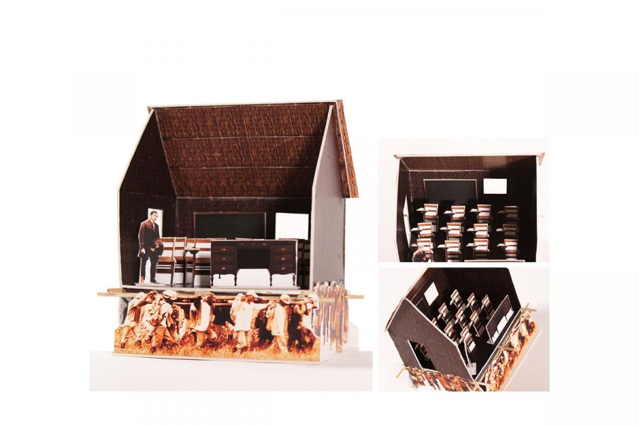 A diorama of a rustic classroom. It features a figure standing beside an old-fashioned desk, surrounded by wooden benches. The structure is barn-like, with straw roofing. Along the base are printed images of people in old-fashioned attire, conveying a historic or educational atmosphere.