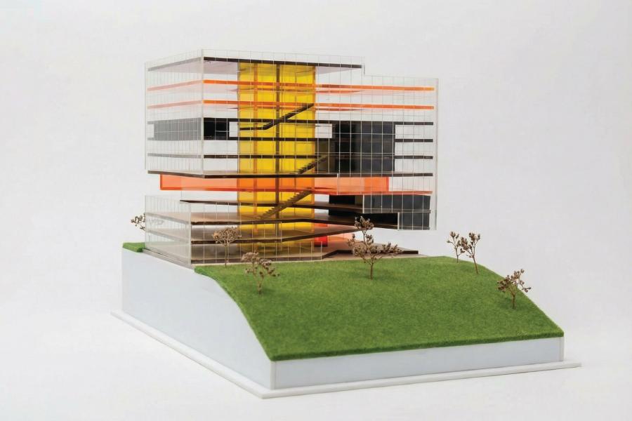 Architectural model of a modern, multi-level building with glass walls accentuated by bright orange and yellow panels, on a grassy base.