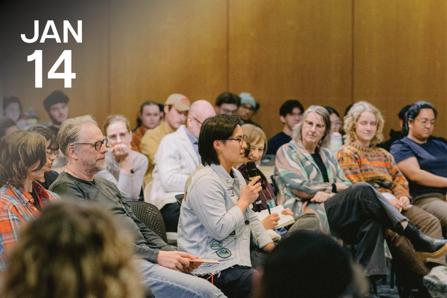 A diverse group of people attentively listens as a person speaks into a microphone in a conference room.