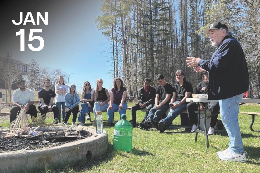A group of people sits outdoors on benches, listening intently to a speaker gesturing beside a table. The setting is grassy with a forest background.
