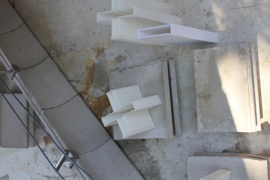 Overhead view of abstract architectural models on a textured concrete surface. The models are white and geometric, casting shadows, with a calm, creative feel.