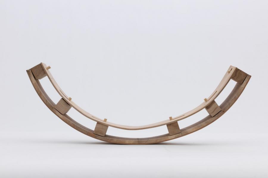 Curved, minimalist wooden structure with a natural finish, featuring a simple arch supported by small, evenly spaced blocks, set against a white background.