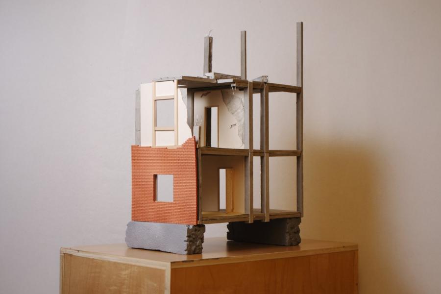 A small architectural model of a partially constructed building, featuring exposed wooden beams, a red brick facade, and an unfinished interior.