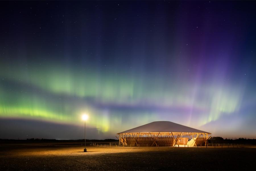A modern wooden pavilion is illuminated under a vibrant aurora borealis, with green and purple hues filling the night sky